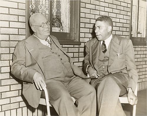 Two men sitting before brick wall with windows and lace curtains