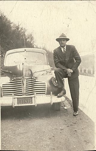 Man wearing suit and hat posed with his foot up on the bumper of a Buick