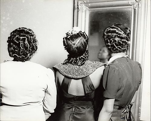 Three women facing a framed mirror modeling hair styles, woman in center wearing dress with exposed back and large collar