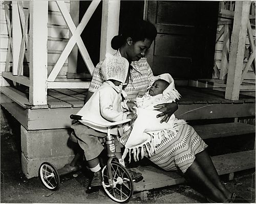 Woman seated on front steps and holding baby, next to child on tricycle