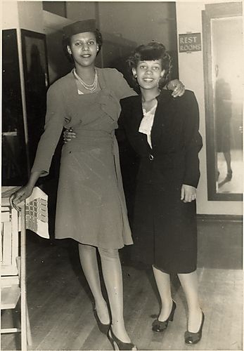 (2 women standing with arms on each other's shoulder and waist)